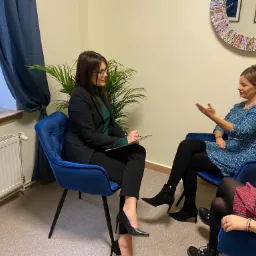 Kobieta w okularach i czarnym garniturze siedzi na niebieskim krześle i robi notatki podczas rozmowy z kobietą w niebieskiej sukience, w tle widać okno z zasłoną i lustro z kolorowych gazet.