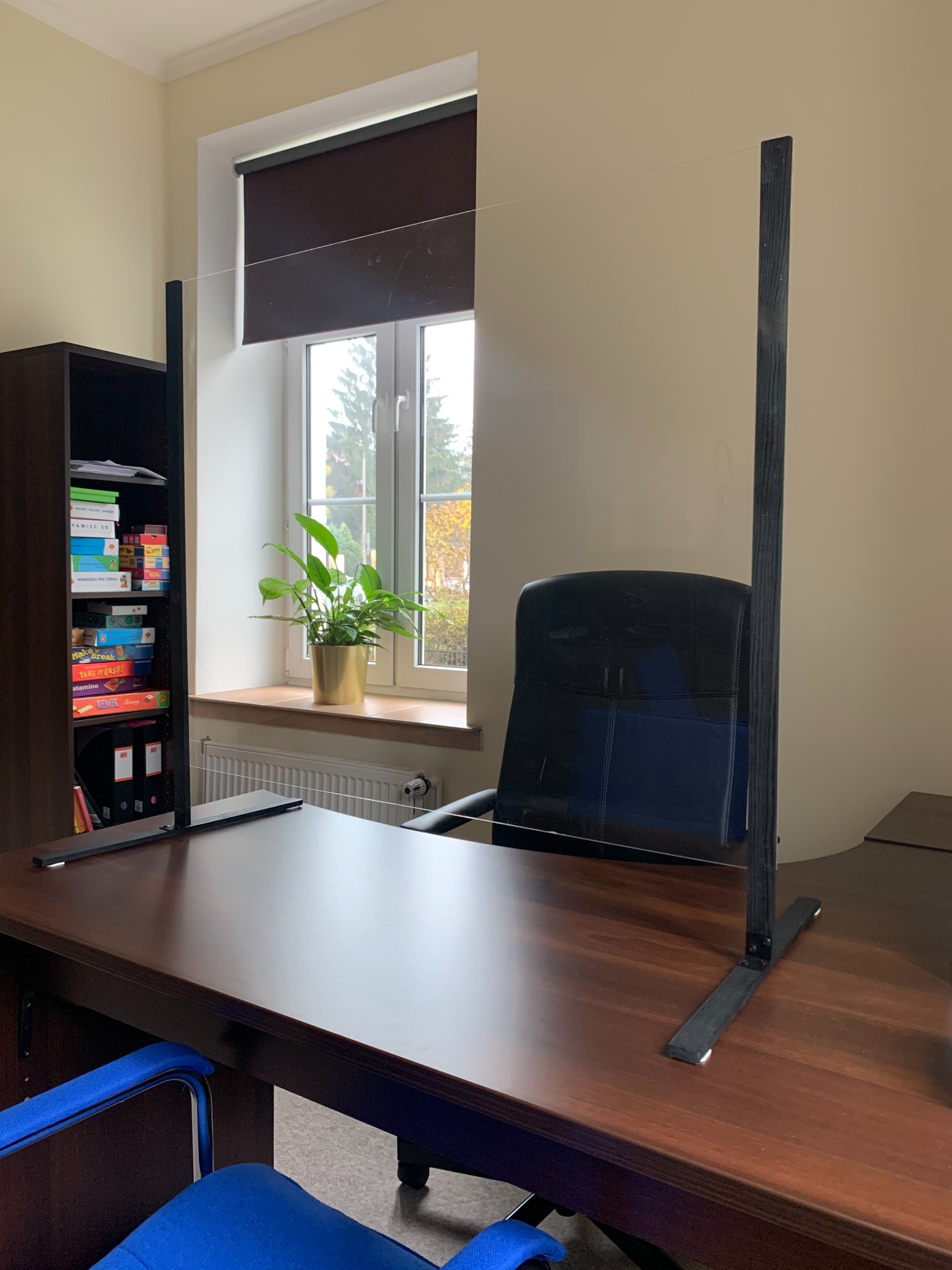 Biurko z przegrodą z pleksi, chroniącą przed rozprzestrzenianiem się zarazków, w tle okno z rośliną doniczkową i regał z książkami.