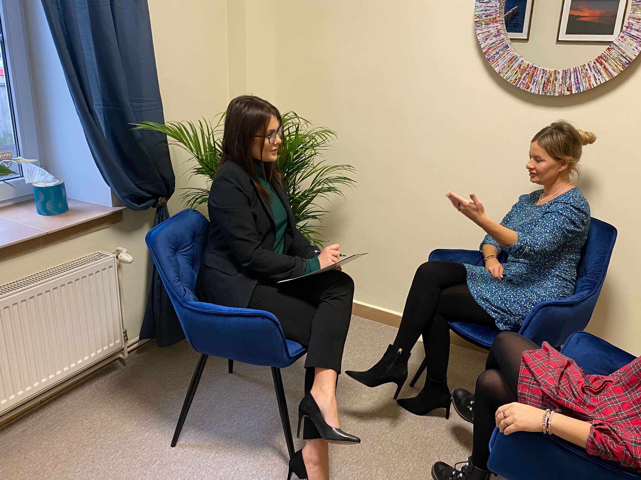 Kobieta w okularach i czarnym garniturze siedzi na niebieskim krześle i robi notatki podczas rozmowy z kobietą w niebieskiej sukience, w tle widać okno z zasłoną i lustro z kolorowych gazet.