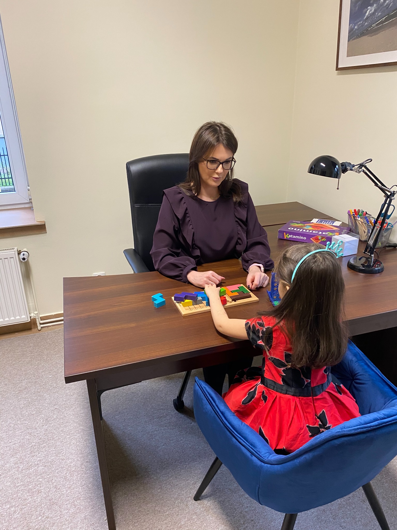 Psycholog dziecięcy z pacjentką układają puzzle geometryczne Katamino na drewnianym biurku w jasnym gabinecie.