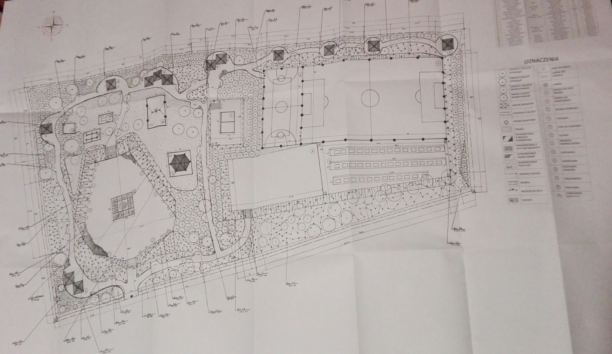 Techniczny rysunek zagospodarowania terenu z boiskami sportowymi, placem zabaw i elementami zieleni, z naniesionymi wymiarami i legendą.