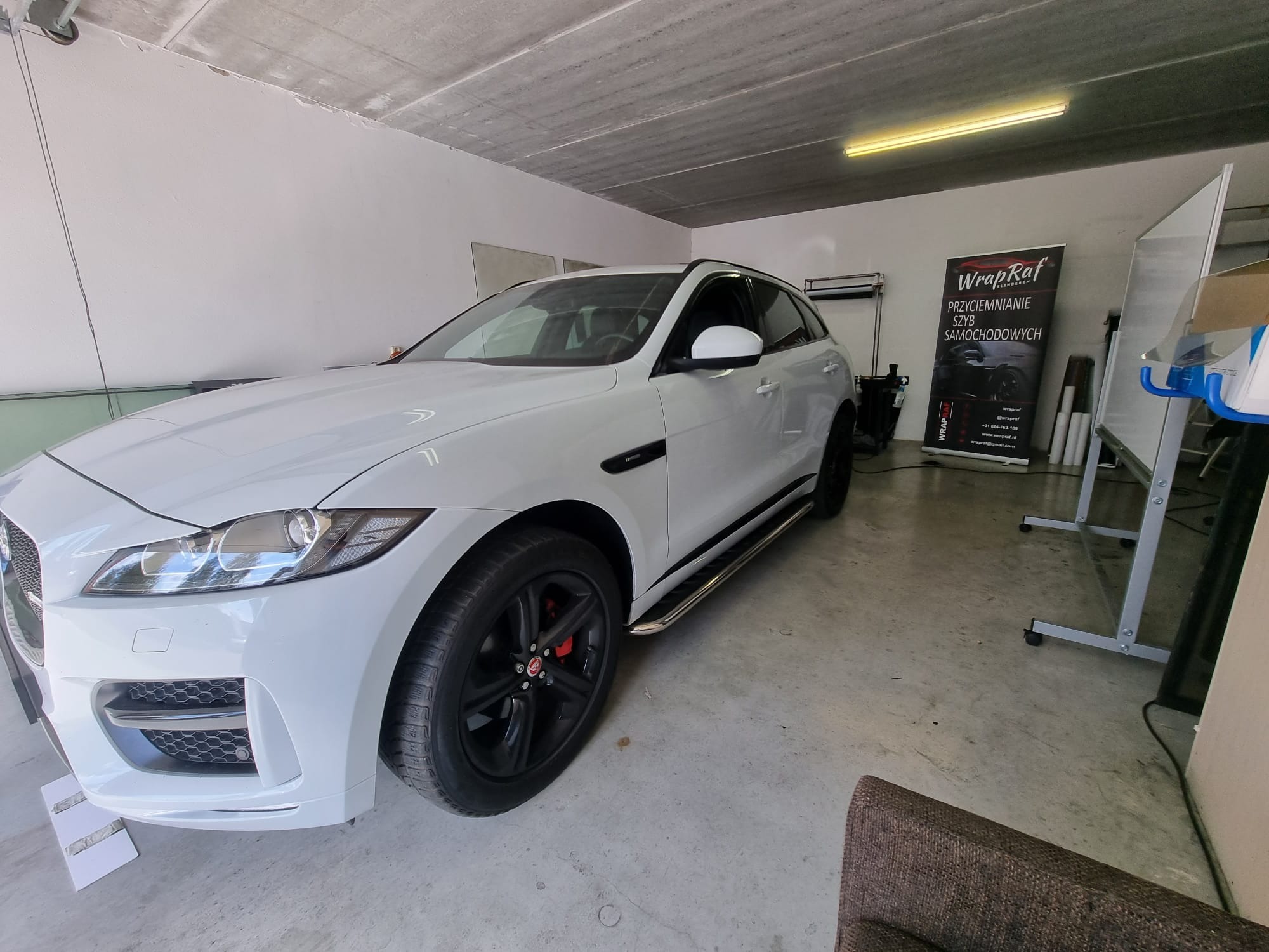 Biały Jaguar F-Pace po usłudze, prawdopodobnie oklejania, w warsztacie z betonową podłogą i ścianami. W tle widoczny banner reklamowy z napisem 'WrapRaf' i informacją o usługach motoryzacyjnych.