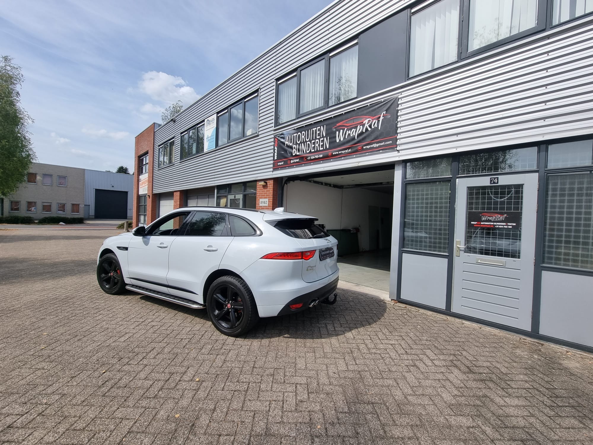 Biały Jaguar F-Pace zaparkowany przed warsztatem z szyldem 'Autoruiten Blinderen WrapRaf', oferującym usługi związane z szybami i oklejaniem samochodów.