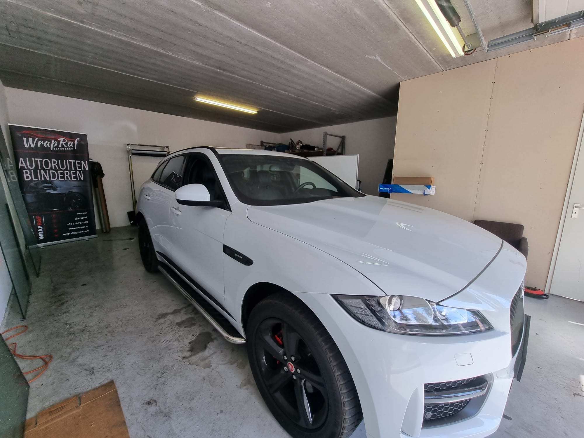 Biały Jaguar F-Pace w garażu, widoczny plakat reklamowy z hasłem 'Autoruiten Blinderen' i logo firmy.