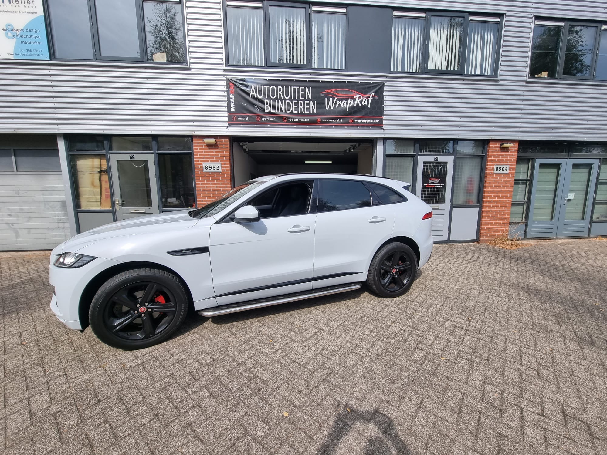 Biały Jaguar F-Pace zaparkowany przed budynkiem z szyldem 'Autoruiten Blinderen Wrap Raf', sugerującym usługi związane z szybami samochodowymi.
