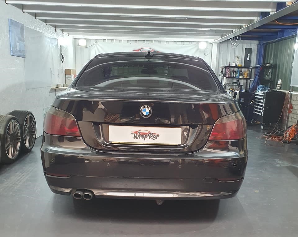 Tył ciemnego BMW serii 5 z przyciemnionymi szybami i tabliczką 'Wrap Rar' w warsztacie, widoczne opony z boku.