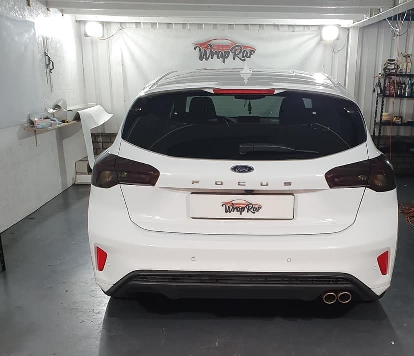 Biały Ford Focus po przyciemnieniu tylnych szyb i oklejeniu klapy bagażnika folią z logo 'Wrap Rar', widoczne ciemne szyby i odblaski światła na lakierze.