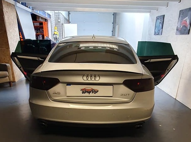 Srebrne Audi A5 z przyciemnionymi tylnymi światłami i szybami, otwarte drzwi ukazują folię na szybach, widoczne logo firmy 'Wrap Rar' na tablicy rejestracyjnej.