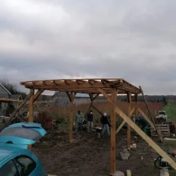 Montaż drewnianej pergoli w ogrodzie: widoczne elementy konstrukcyjne, ekipa montażowa oraz otwarty bagażnik niebieskiego samochodu osobowego w tle wiejskiego krajobrazu.