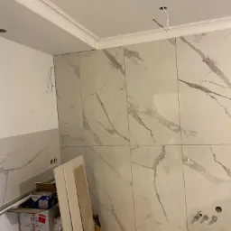 Montaż płytek imitujących marmur w łazience w Tczewie, widoczne przygotowane otwory na armaturę, niezabezpieczone przewody elektryczne oraz kartony po materiałach budowlanych.