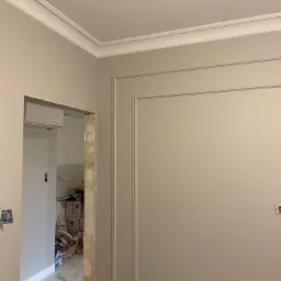 Ściana w jasnym odcieniu z dekoracyjnymi listwami i sztukaterią, widoczne przygotowania do montażu klimatyzacji i elementy wykończeniowe w przejściu do innego pomieszczenia.