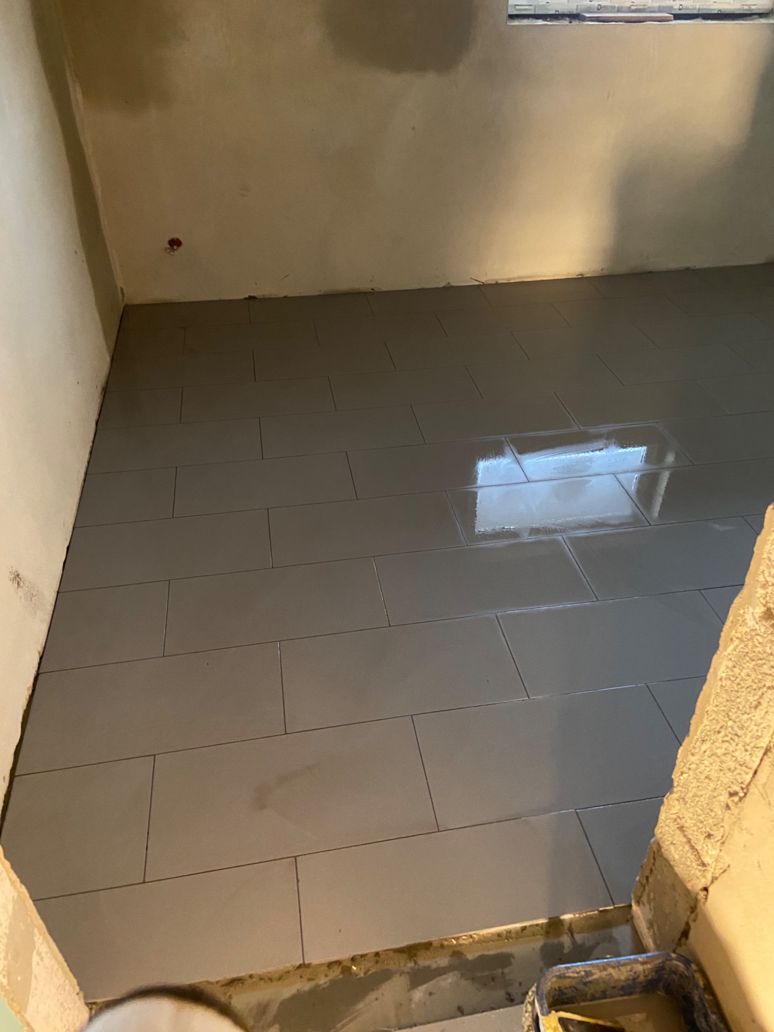 Ułożone, prostokątne, szare płytki podłogowe w pokoju z surowymi ścianami i widocznym odbiciem okna, z widocznym wiaderkiem na fugę w rogu.