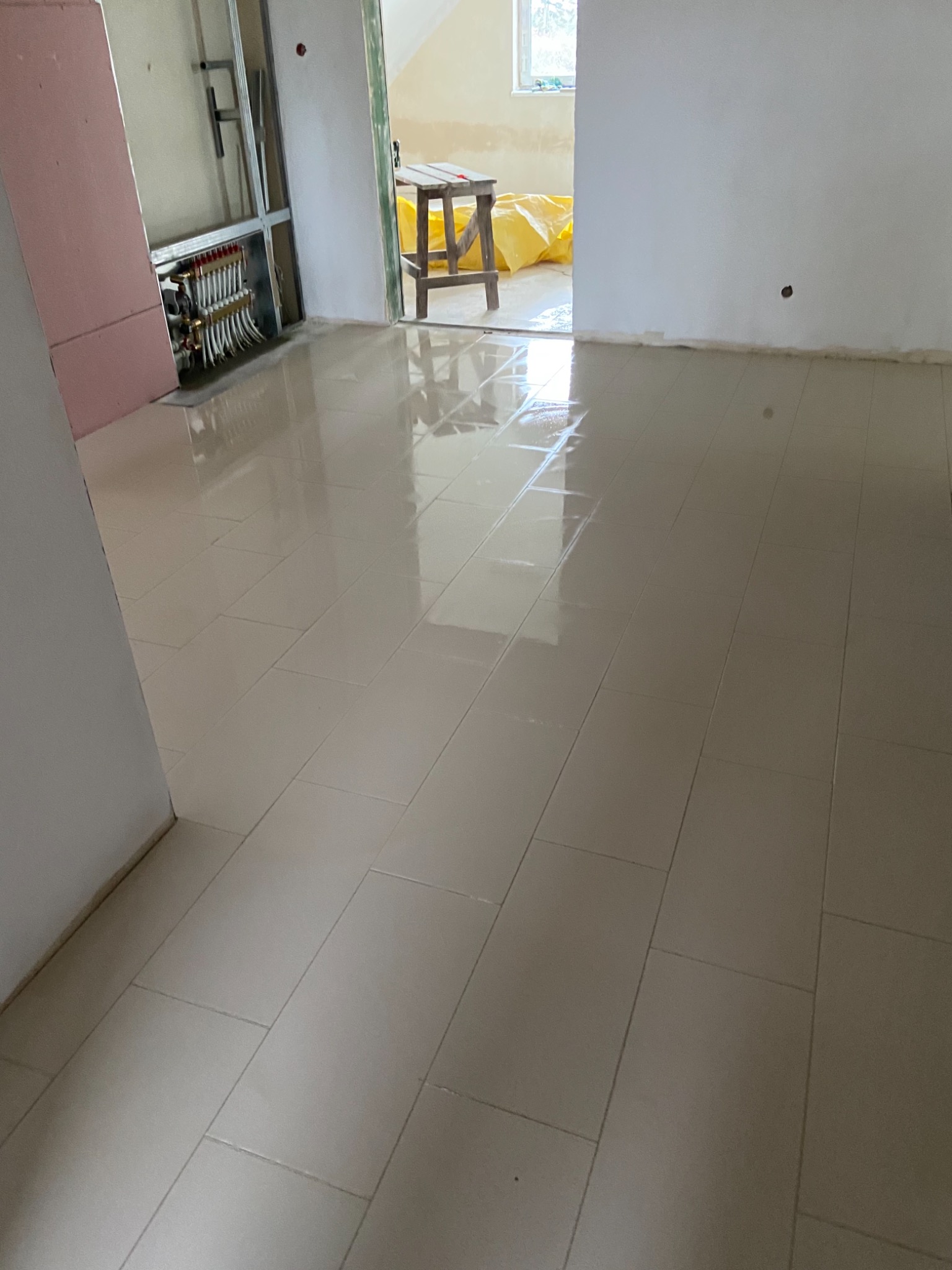 Błyszcząca, beżowa podłoga z płytek ceramicznych ułożonych w prostokątnym wzorze, widoczne elementy instalacji grzewczej podłogowej i fragment drabiny w tle, pomieszczenie w trakcie wykańczania.