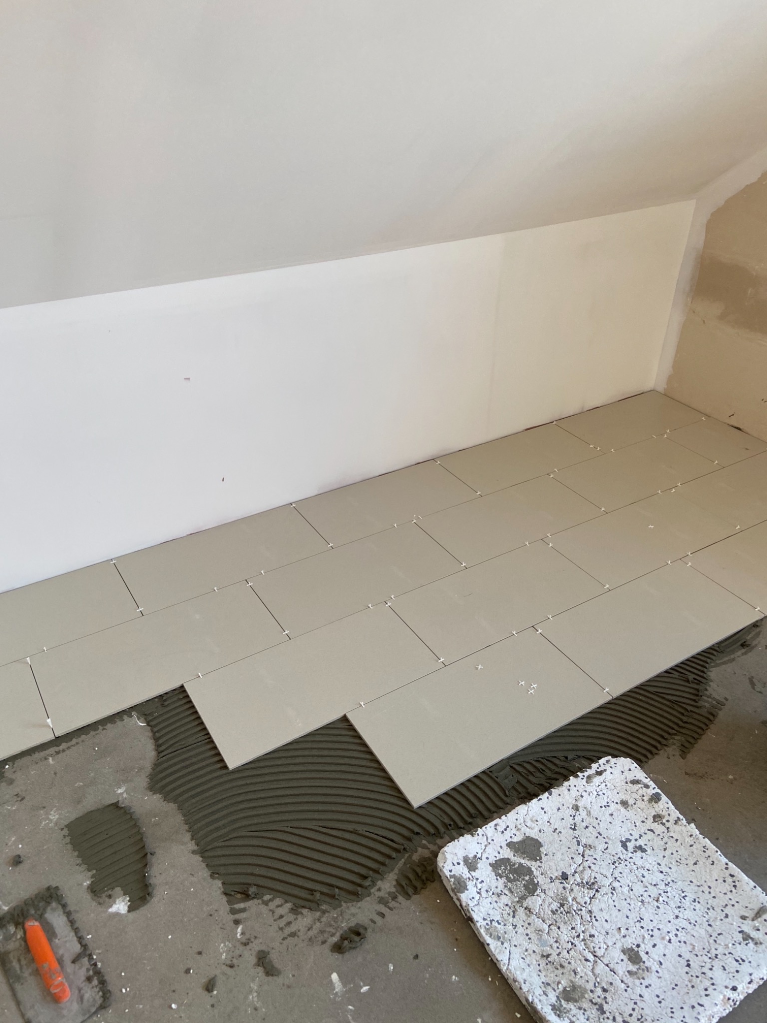 Układanie jasnych, prostokątnych płytek podłogowych w skos na poddaszu, widoczna warstwa kleju, krzyżyki dystansowe, brudna paca i surowe ściany.