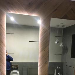 Mc Rem-House Remonty-Wykończenie Wnętrz - Nowoczesna łazienka z dwoma lustrami: jedno podświetlane LED, drugie z widocznym odbiciem armatury i lamp wiszących, ściany wykończone drewnopodobnymi płytkami ułożonymi w jodełkę.