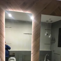 Nowoczesna łazienka z dwoma lustrami: jedno podświetlane LED, drugie z widocznym odbiciem armatury i lamp wiszących, ściany wykończone drewnopodobnymi płytkami ułożonymi w jodełkę.