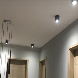Mc Rem-House Remonty-Wykończenie Wnętrz - Nowoczesne oświetlenie sufitowe: czarne reflektory punktowe i lampa wisząca z długimi przewodami, jasne ściany i drewniane drzwi w korytarzu.