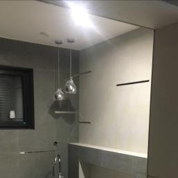 Mc Rem-House Remonty-Wykończenie Wnętrz - Szara łazienka w trakcie wykańczania z wiszącymi lampami w folii ochronnej, widoczne płytki, okno z żaluzjami i niedokończone wykończenie ściany przy wejściu.