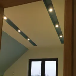 Ukośny sufit poddasza z wbudowanymi, punktowymi lampami LED w niebieskich wnękach, widok zza drewnianej framugi drzwi na okno z widokiem na zewnątrz.