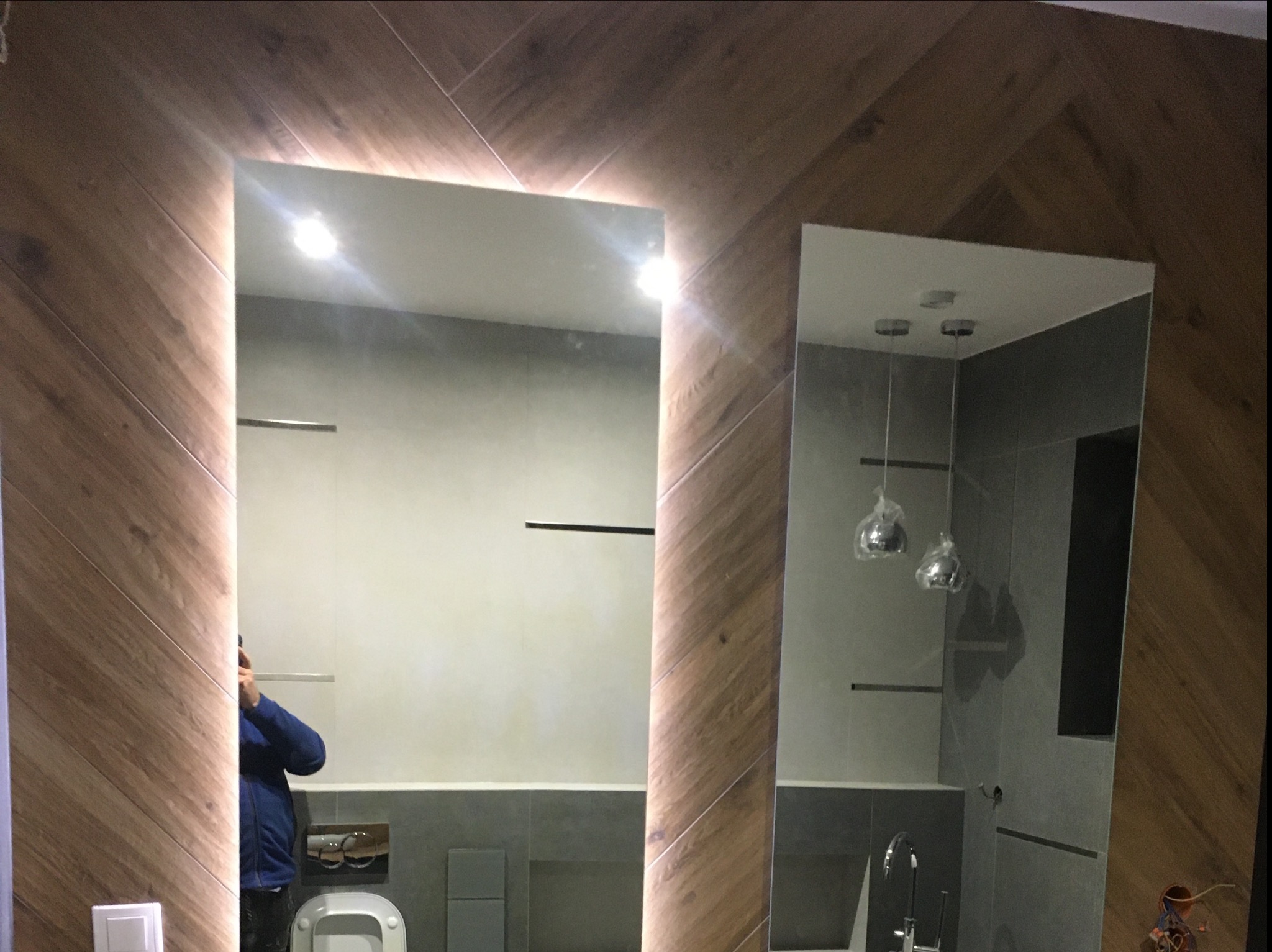 Nowoczesna łazienka z dwoma lustrami: jedno podświetlane LED, drugie z widocznym odbiciem armatury i lamp wiszących, ściany wykończone drewnopodobnymi płytkami ułożonymi w jodełkę.