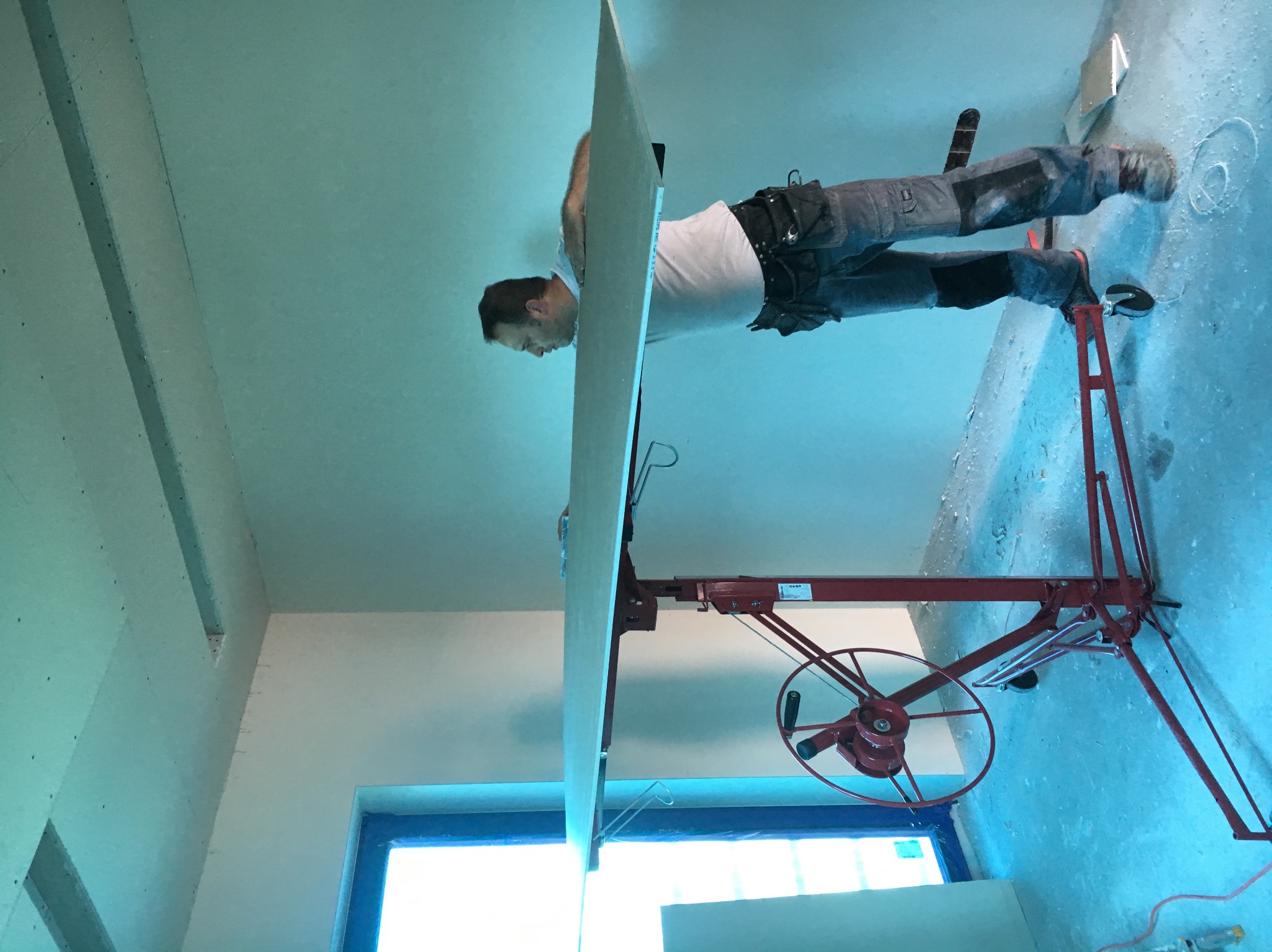 Pracownik montuje płytę gipsowo-kartonową na suficie za pomocą podnośnika; widoczne fragmenty oświetlenia i okna zabezpieczonego folią.