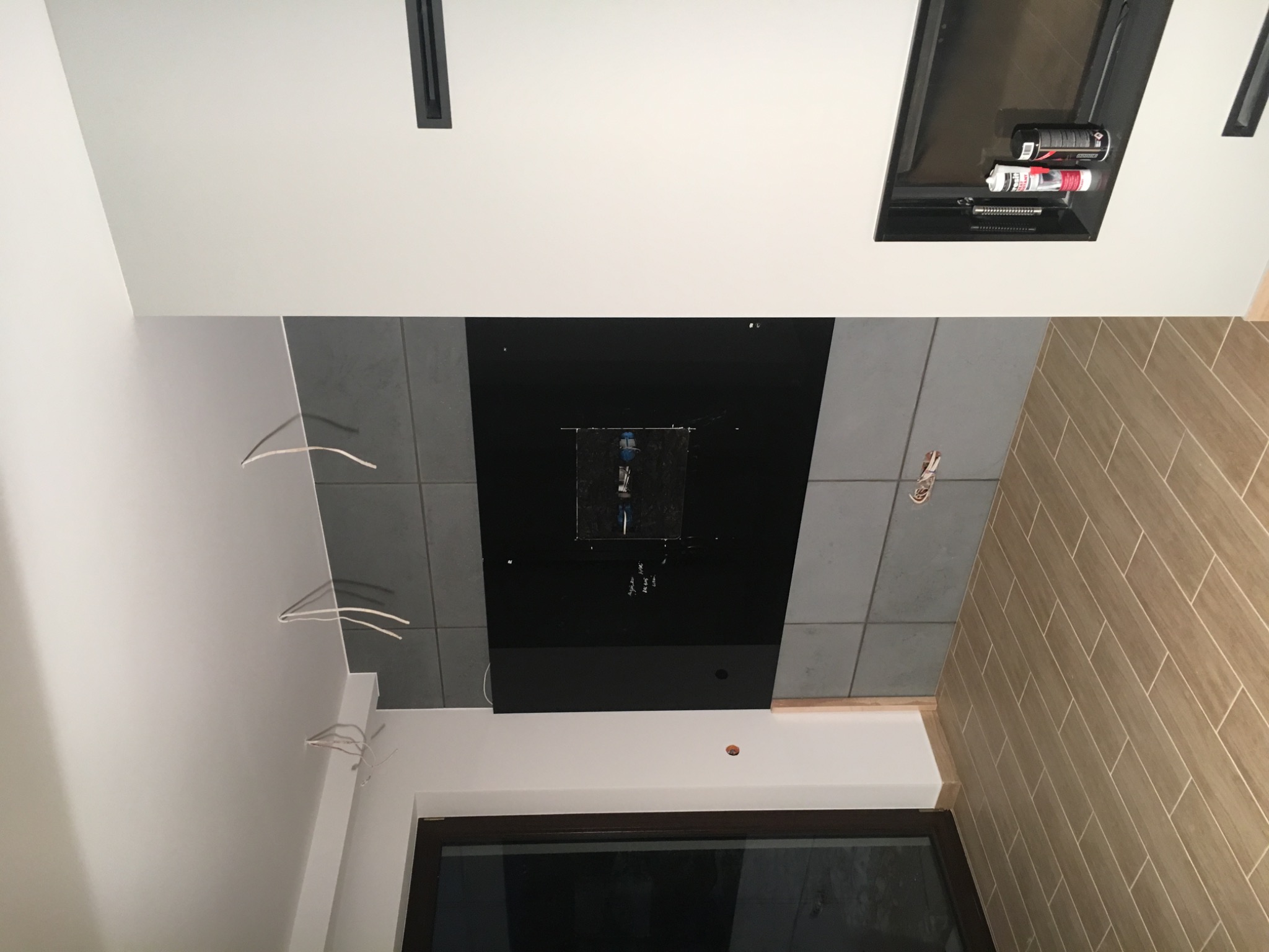 Częściowo wykończona ściana z szarymi płytkami i czarnym panelem, widoczne wystające kable elektryczne, zabudowa z narzędziami i materiałami montażowymi oraz fragment ściany wyłożonej płytkami...