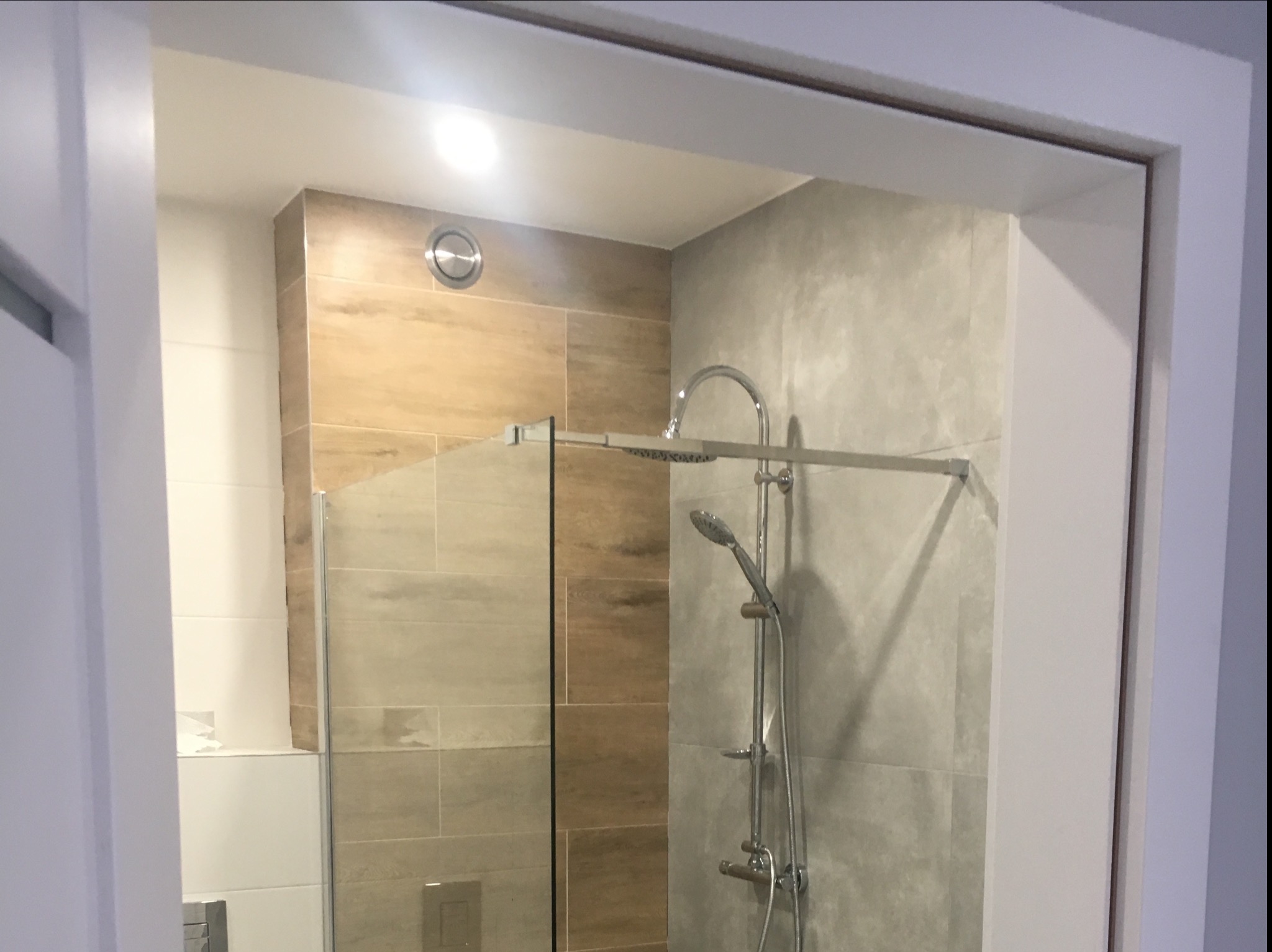 Nowoczesna kabina prysznicowa walk-in z deszczownicą i baterią termostatyczną, wykończona płytkami w odcieniach beżu i szarości, widoczna przez framugę drzwi.