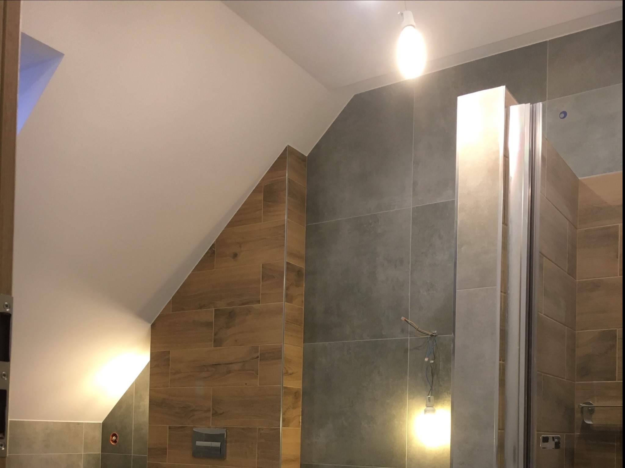 Ukośna łazienka z szarymi i drewnopodobnymi płytkami, widoczne niedokończone podłączenie oświetlenia.