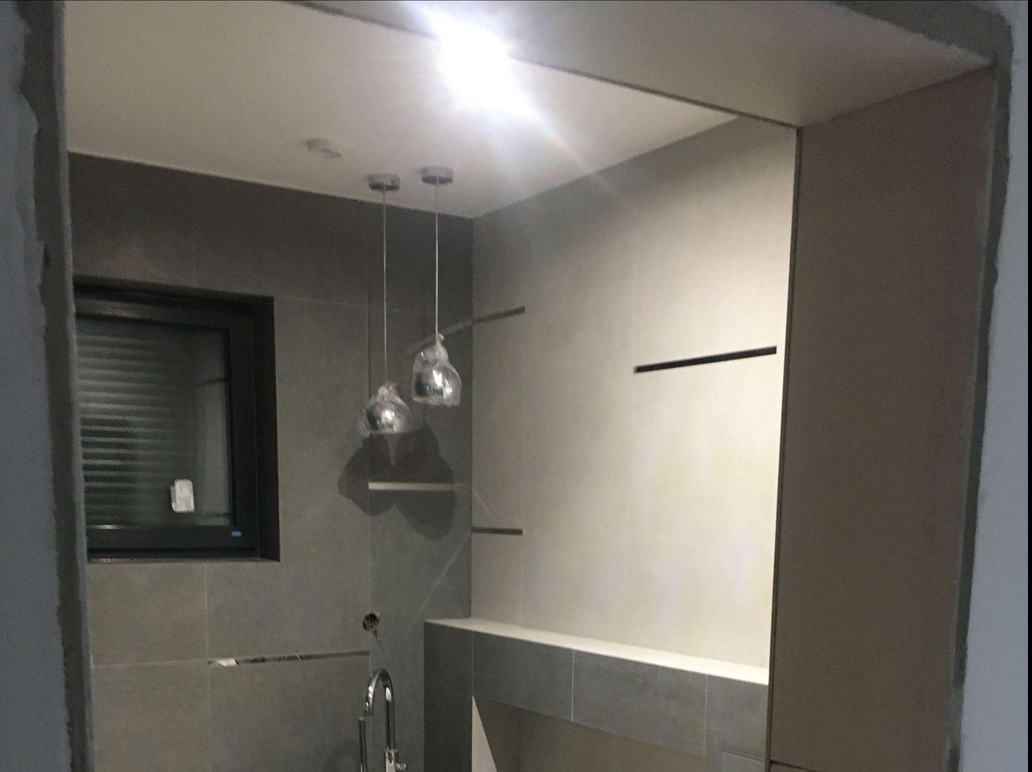Szara łazienka w trakcie wykańczania z wiszącymi lampami w folii ochronnej, widoczne płytki, okno z żaluzjami i niedokończone wykończenie ściany przy wejściu.