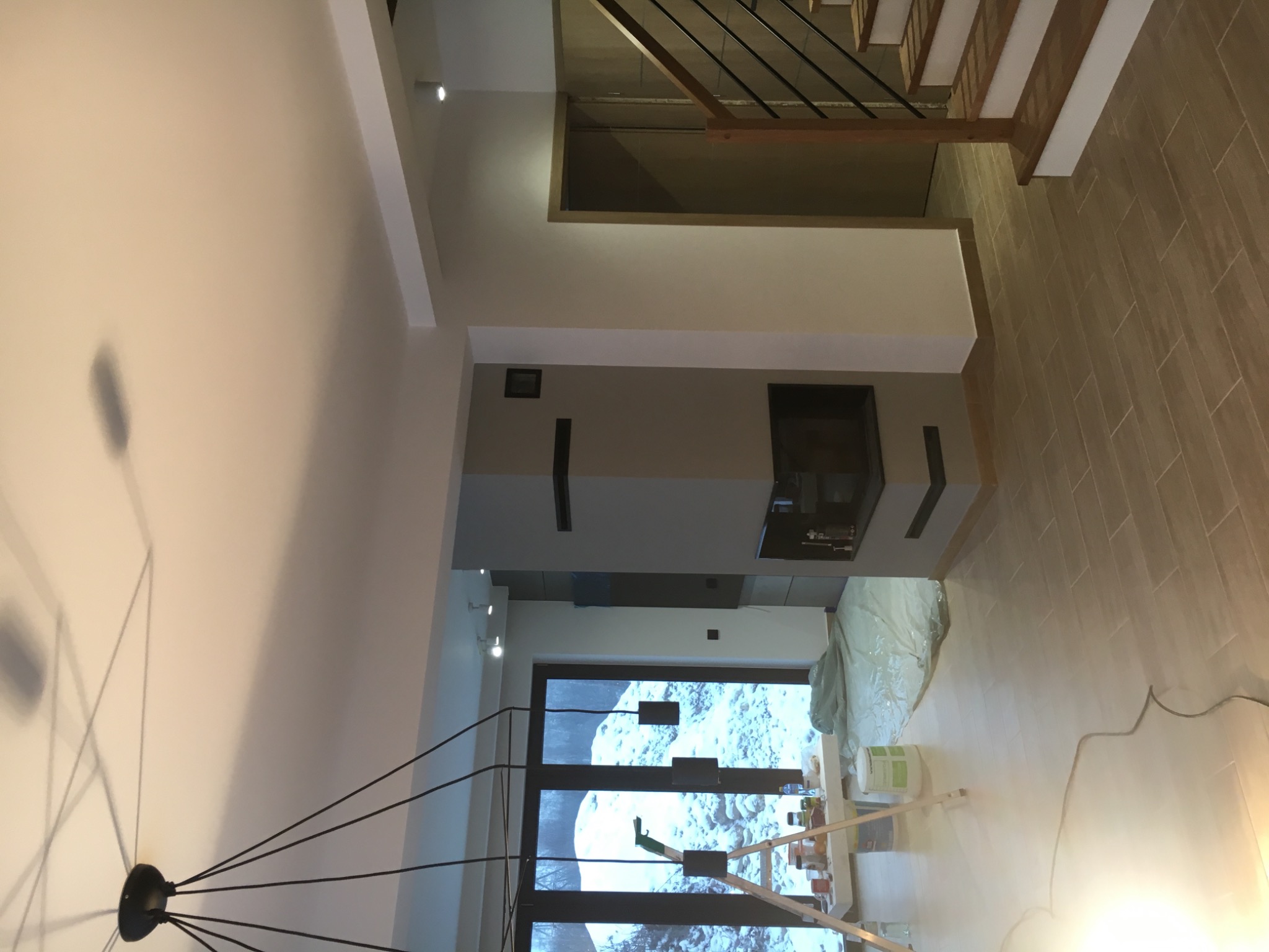 Wnętrze w trakcie remontu: widok na kominek z szarej płyty, zabezpieczone folią podłogi i schody z metalową balustradą; widoczna drabina malarska i narzędzia malarskie.