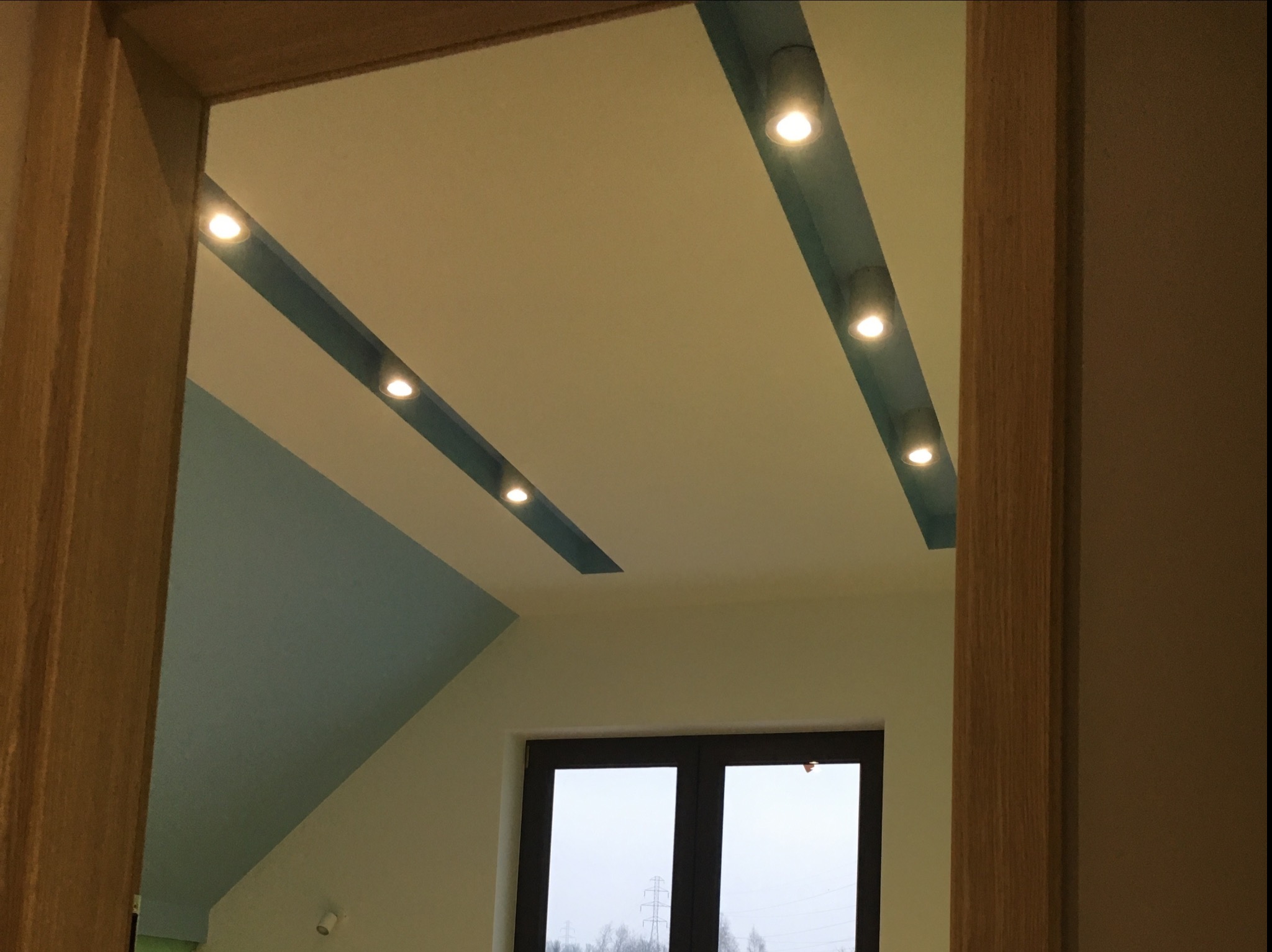 Ukośny sufit poddasza z wbudowanymi, punktowymi lampami LED w niebieskich wnękach, widok zza drewnianej framugi drzwi na okno z widokiem na zewnątrz.