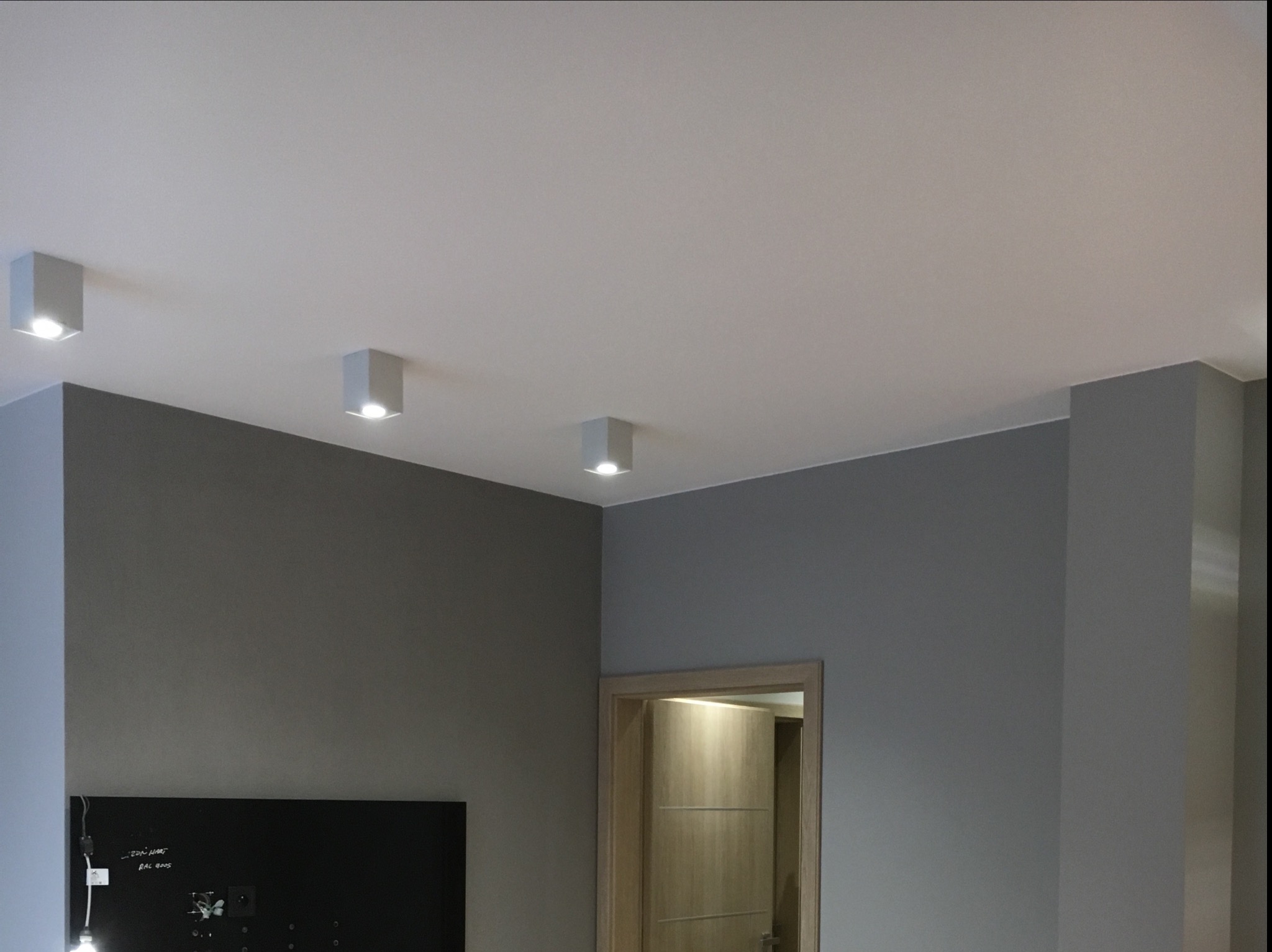 Szare wnętrze pokoju z trzema kwadratowymi lampami sufitowymi LED, ciemną ścianą z tablicą i widocznymi przewodami, oraz otwartymi drzwiami do innego pomieszczenia z drewnianymi drzwiami.
