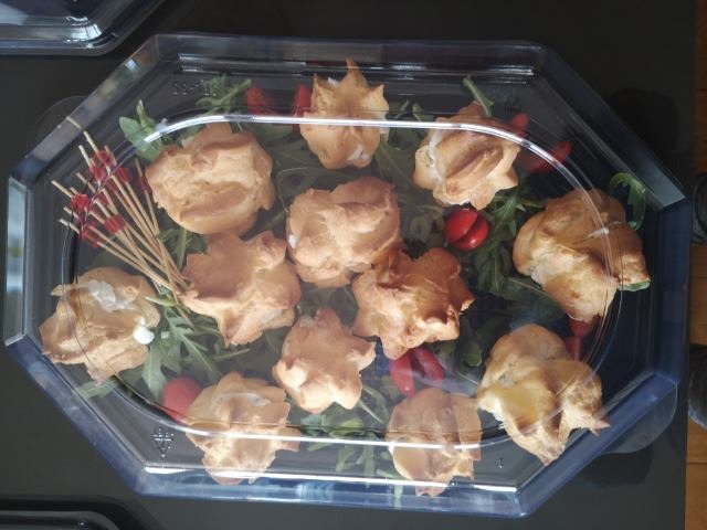Profiterolki z kremem w przezroczystym opakowaniu cateringowym, ułożone na rukoli i pomidorkach koktajlowych, z dekoracyjnymi wykałaczkami.