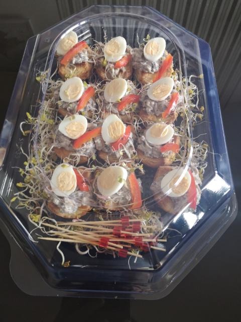 Przezroczyste opakowanie z przekąskami: kanapeczki z pastą, jajkiem przepiórczym, czerwoną papryką i kiełkami rzodkiewki, obok ułożone drewniane wykałaczki z czerwonymi końcówkami.