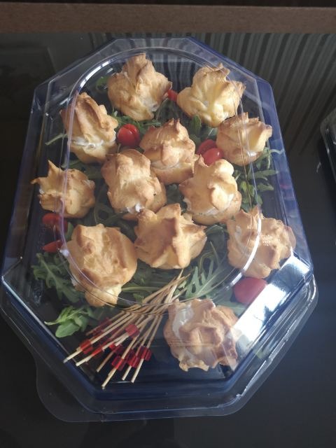 Patera z przezroczystego plastiku wypełniona ptysiami z kremem, rukolą i pomidorkami koktajlowymi, uzupełniona o dekoracyjne wykałaczki z czerwonymi końcówkami.