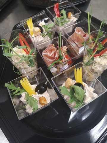 Kreatywnie podane przystawki w przezroczystych naczyniach na czarnej tacy: sałatka z jajkiem, szynka parmeńska, kolorowe papryki, rukola i oliwki.