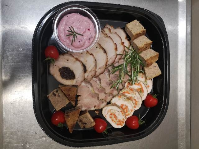 Czarny plastikowy półmisek z selekcją wędlin, pasztetów, różowego sosu z rozmarynem i pomidorków koktajlowych, ułożonych na metalowym blacie.