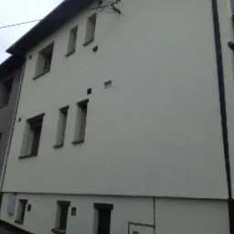 Dwukondygnacyjny dom z elewacją w odcieniach bieli i szarości, widok z zewnątrz, z widocznymi oknami o różnych rozmiarach i kształtach oraz elementami dekoracyjnymi.