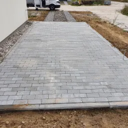 Ułożona szara kostka brukowa na podjeździe, obrzeża z kostki, w tle biały bus i fragment budynku.