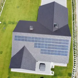 Widok z lotu ptaka na dom z ciemnoszarą dachówką, na której zamontowano panele słoneczne; zadbany trawnik i ścieżka wzdłuż ogrodzenia.