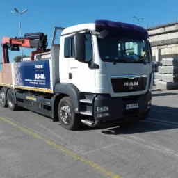 Ciężarówka MAN z dźwigiem załadowana kostką brukową Polbruk AD-OL na placu, widoczne palety z kostką w tle.