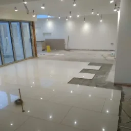 Układanie jasnych, prostokątnych płytek podłogowych w przestronnym, jasnym pomieszczeniu z dużymi przesuwnymi drzwiami balkonowymi i licznymi punktowymi lampami sufitowymi; widoczne niedokończone...