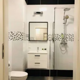 Nowoczesna łazienka z białymi płytkami, czarnymi akcentami i szklanym prysznicem; widoczna umywalka z szafką, lustro w czarnej ramie i toaleta.