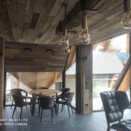 Rustykalne wnętrze z drewnianym sufitem i ścianami, okrągły stół z krzesłami i widokiem na zewnątrz przez duże okna.