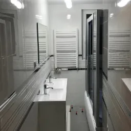 Wąska, jasna łazienka z dwoma umywalkami na blacie, grzejnikiem drabinkowym, kabiną prysznicową i poziomowaniem płytek podłogowych przy użyciu klipsów i klinów.