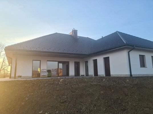 Nowo wybudowany, parterowy dom z ciemną dachówką i białymi ścianami, widoczny z poziomu gruntu, z niewielkim nasypem ziemi przed budynkiem i materiałami budowlanymi przy oknie.