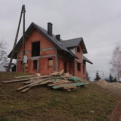 Surowy stan domu z czerwonej cegły, widoczne deski na ziemi, brak okien i drzwi, dach pokryty czarną dachówką, szare niebo w tle.
