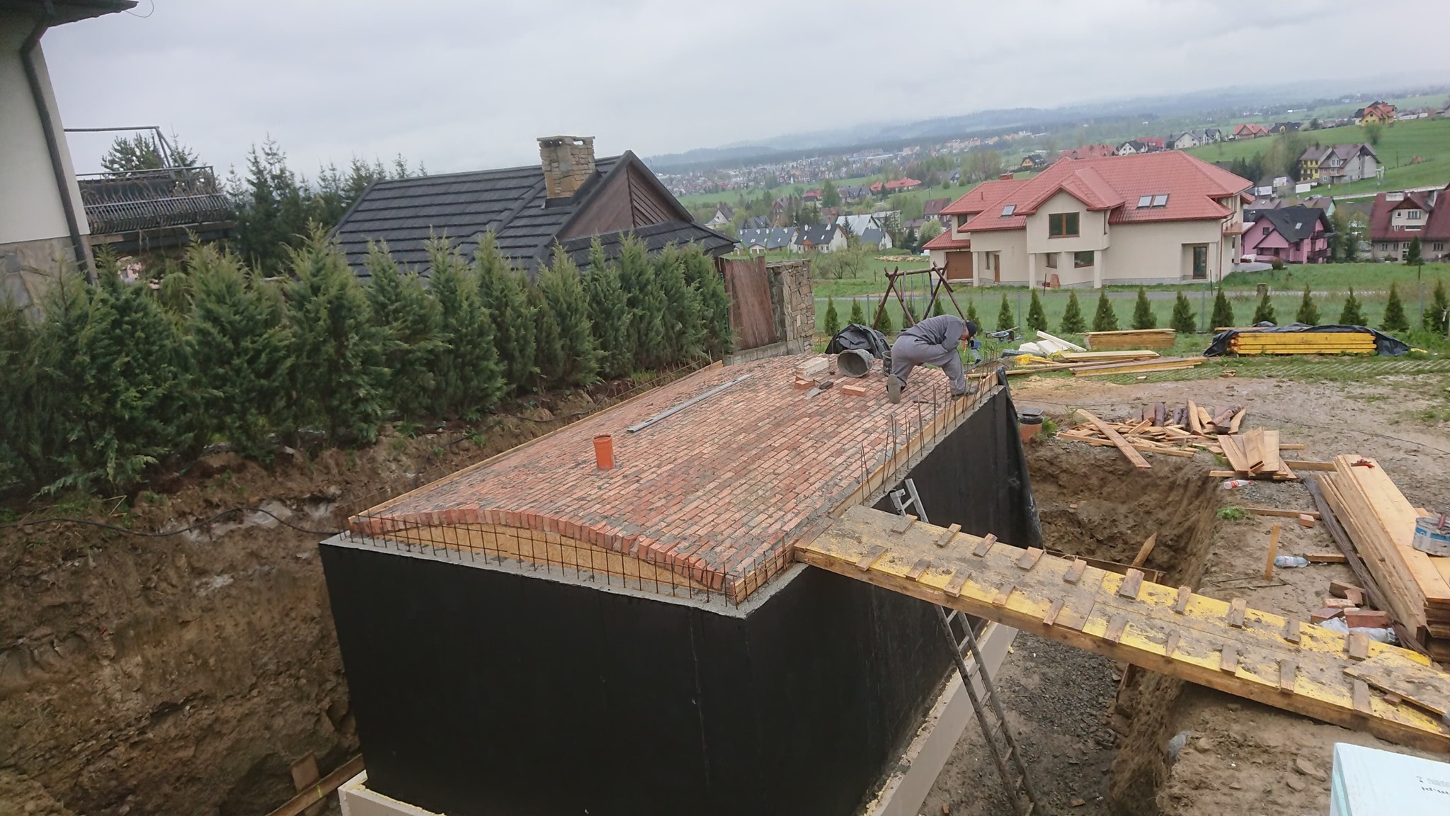 Budowa piwnicy: Widok z góry na zbrojoną konstrukcję z ceglanym stropem, zabezpieczoną czarną izolacją, z pracownikiem w szarym kombinezonie układającym cegły na tle krajobrazu z domami i wzgórzami.