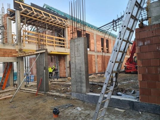 Wznoszony dwukondygnacyjny budynek z czerwonej cegły i betonu, widoczne szalunki stropowe, zbrojenie pionowe i drabina oparta o ścianę, w tle czerwony dźwig i robotnicy w żółtych kamizelkach.