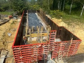 Wykop pod fundamenty z zamontowanym szalunkiem systemowym w kolorze czerwonym, widoczne zbrojenie i folia izolacyjna wewnątrz, teren zielony w tle.